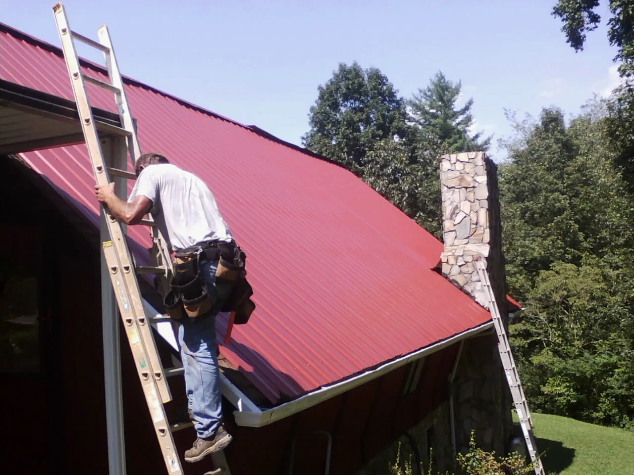 Roofer on a ladder beside a red metal roof and stone chimney — Soffit & Fascia Repair by Roofing Brothers in Lithopolis